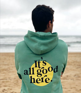 Sudadera unisex en color verde con capucha y mensaje "it's all good here" en la parte trasera y smilie en la delantera. Fabricada con algodón orgánico y poliéster reciclado. Sudadera sostenible de Lorem Ipsum Brand